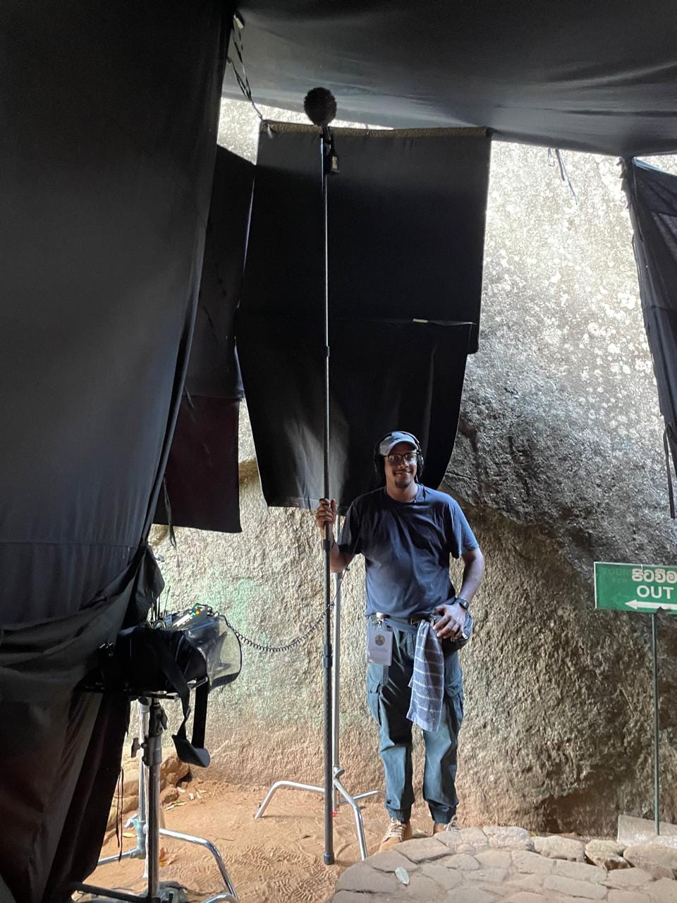 Chandana operating a boom microphone on a film location beneath draped black flags and rock